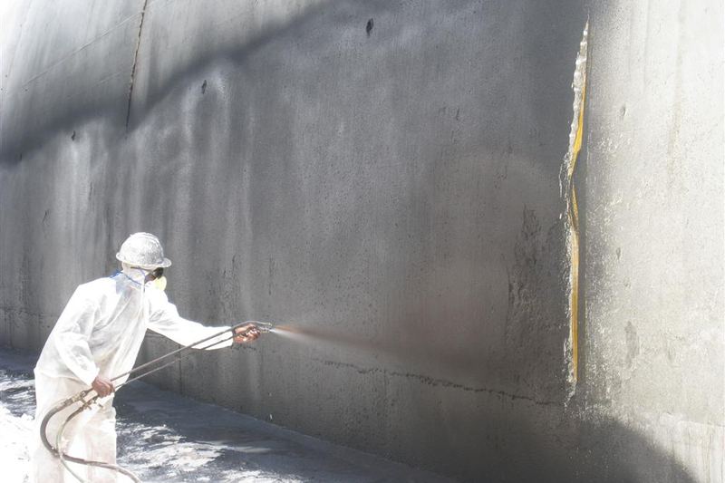 Worker spray-applying waterproofing material to an exterior concrete wall
